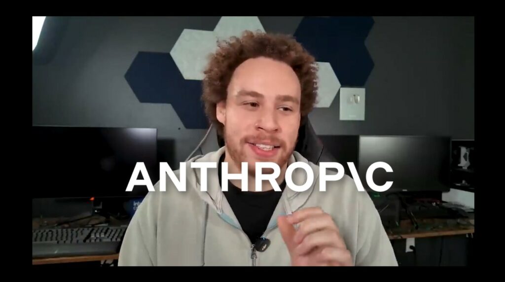 Primo piano di Marcus Hutchins davanti a una postazione da lavoro, con sfondo composto da pannelli esagonali decorativi e monitor spenti. Indossa una felpa chiara e un microfono clip. Sopra l’immagine è sovrapposta la scritta grande “ANTHROPIC”, suggerendo un contesto legato all’intelligenza artificiale e alla tecnologia.