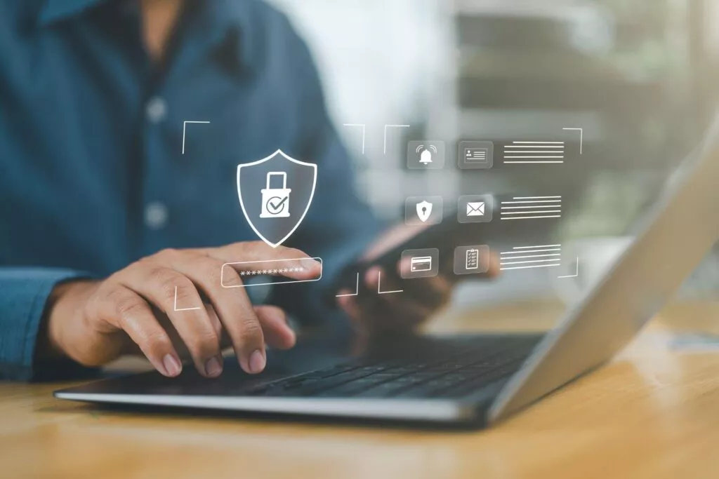Uomo che usa un computer portatile e uno smartphone con icone grafiche sovrapposte riguardanti la sicurezza informatica e la privacy.