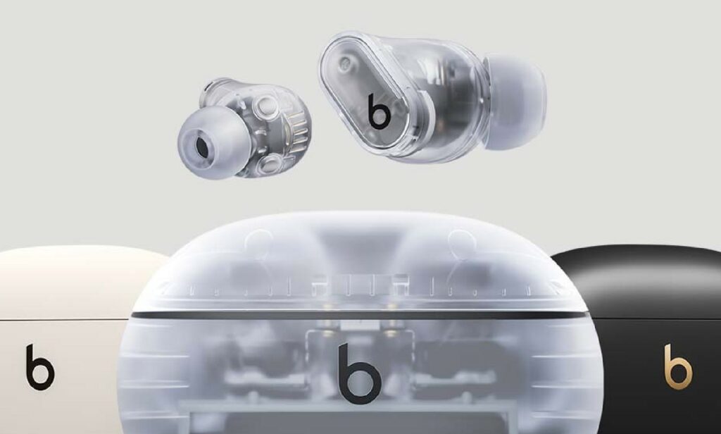 Questa immagine mostra un primo piano promozionale di un set di auricolari true wireless firmati Beats, caratterizzati da un design moderno e da diverse varianti di colore. Nella parte superiore dell'inquadratura fluttuano i due auricolari in una suggestiva versione completamente trasparente. Questa finitura permette di intravedere chiaramente i circuiti interni, i componenti dorati, i magneti e la struttura tecnologica sottostante, conferendo al prodotto un look futuristico e minimalista di ispirazione industriale. Sulla superficie esterna di ciascun auricolare spicca il celebre logo con la lettera "b" di colore nero. Gli auricolari sono completati da gommini in silicone grigio chiaro semitrasparente. Nella metà inferiore dell'immagine sono parzialmente visibili tre diverse custodie di ricarica disposte una accanto all'altra, che suggeriscono le opzioni cromatiche disponibili per questo modello. Al centro domina la custodia corrispondente alla versione trasparente degli auricolari, realizzata con una plastica opaca e satinata che lascia intravedere l'alloggiamento interno e i contatti metallici per la ricarica, con il logo nero posizionato frontalmente. Alla sua sinistra si scorge una parte di una custodia di colore avorio o crema chiaro con il logo nero, mentre sulla destra emerge una porzione di una variante nera opaca, impreziosita da un logo Beats di un elegante colore oro o bronzo. Lo sfondo dell'intera composizione è completamente neutro, caratterizzato da una tonalità grigio chiaro uniforme che fa risaltare i dettagli del design, i riflessi sulle superfici plastiche e i loghi del brand.