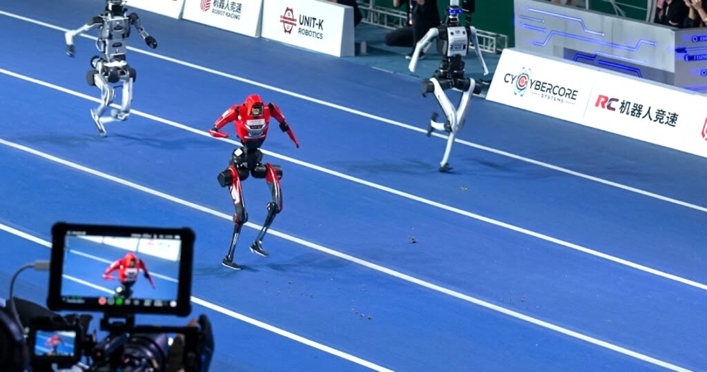Atleti in rosso e bianco su una pista, con robot che corrono in primo piano, durante una competizione sportiva.